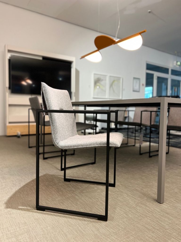 Modern office meeting area with gray upholstered chair and metal frame beside a long table; contemporary pendant light overhead.