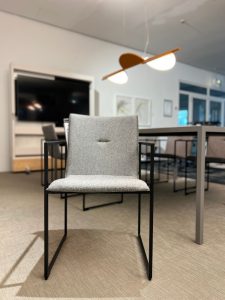 Grey fabric conference chair with a black metal frame in a modern meeting room, foreground with blurred table and pendant light in the background.