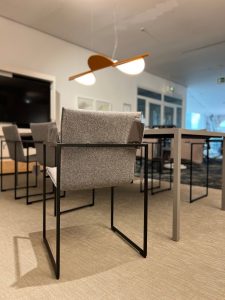 Empty modern conference room with gray upholstered chairs and metal-frame desks under a wooden two-panel pendant light.