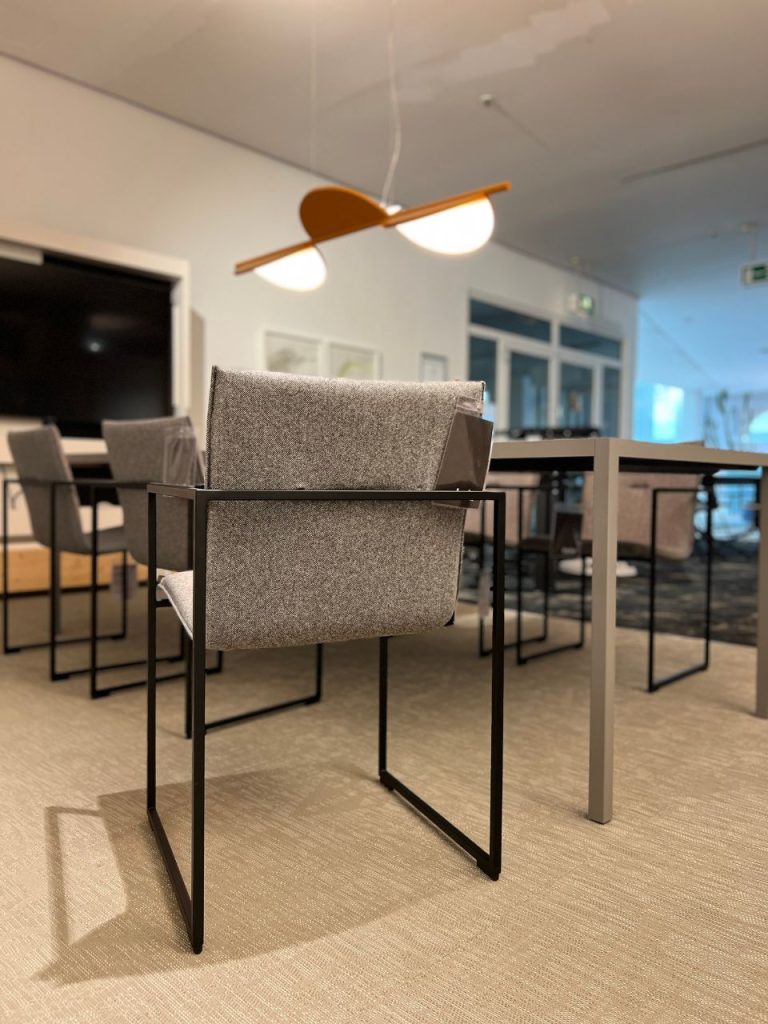 Empty modern conference room with gray upholstered chairs and metal-frame desks under a wooden two-panel pendant light.