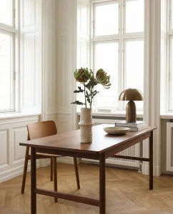 Wood dining table in a sunlit, white-paneled room with a tall vases of greenery, a brass lamp, and a small bowl on stacked books beside it