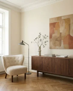 Minimalist living room corner: a cream boucle armchair beside a black floor lamp, a wooden sideboard with books and a vase of branches, and an abstract beige-brown painting on the wall by a large window.