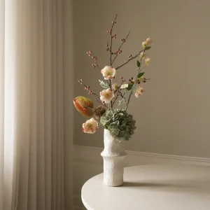 Flower arrangement in a white textured vase on a round white table, with beige curtains in the background.