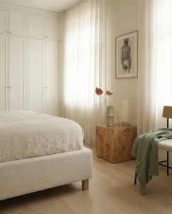 Neutral bedroom with a white built-in wardrobe, a light linen bed, and a wooden cube side table by sheer curtains.