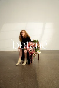 Woman in a black dress and beige boots sits beside a chair overflowing with pink and white flowers; brand name 'boltené' overlays the image.