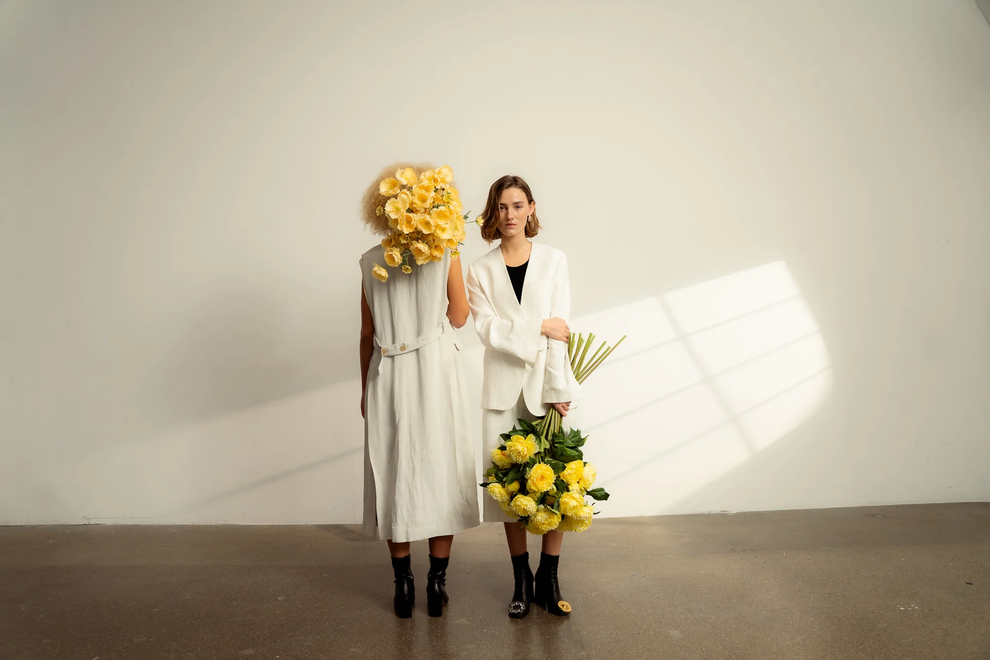 Two women in white outfits stand against a pale wall; the left person has a large yellow flower bouquet obscuring their head, while the right holds a cluster of yellow flowers and looks at the camera.