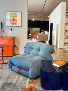 Blue tufted lounge chair beside a glossy dark blue coffee table with an orange notebook in a modern furniture showroom.