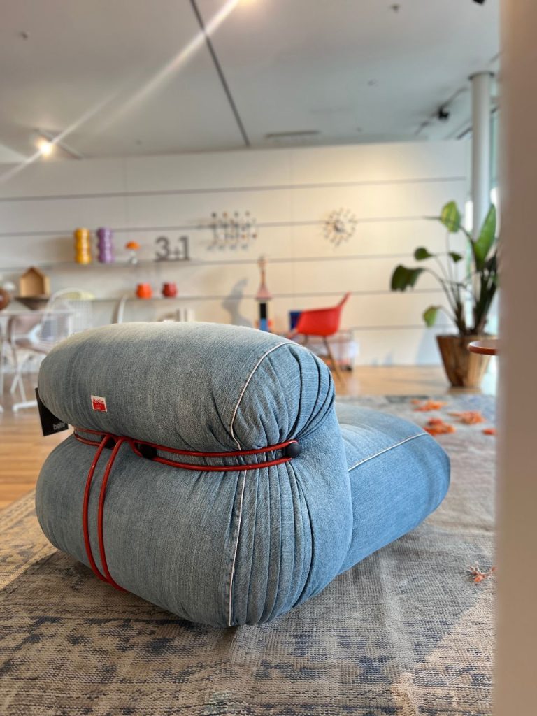Blue denim pouf with a red cord wrapped around it sits on a patterned rug in a bright, modern showroom.