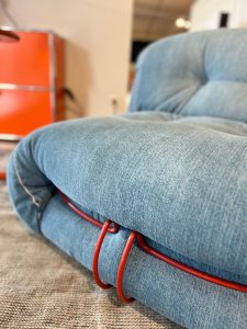 Blue denim upholstered chair/sofa cushion with bright red cables looped around the edge, resting on a beige carpet.