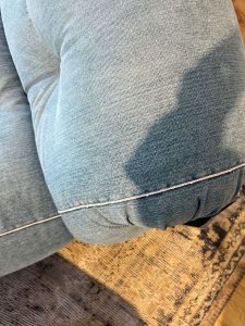 Close-up of a light blue denim sofa armrest with white piping, resting on a patterned rug nearby.