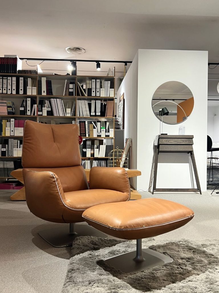 Brown leather lounge chair with matching ottoman on a shaggy gray rug, in a showroom with a shelving wall of binders and books behind it and a white partition with a circular mirror to the right.