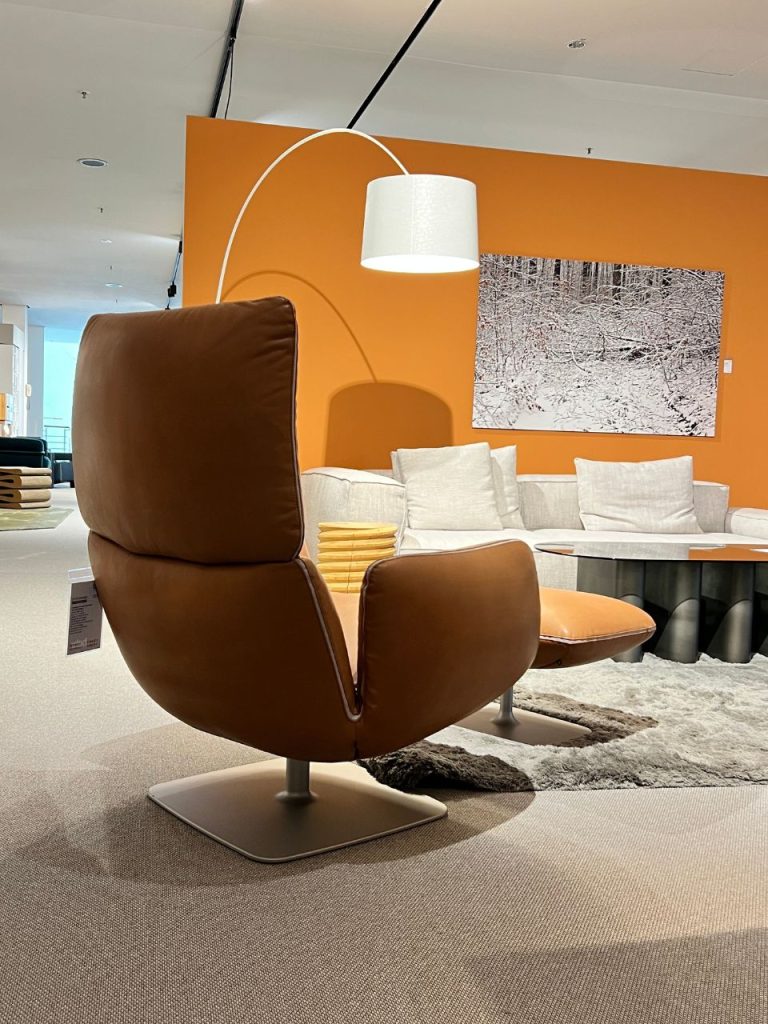Brown leather high-back lounge chair with matching ottoman in a showroom, white sofa, orange accent wall, arc floor lamp, and a snowy forest artwork on the wall.