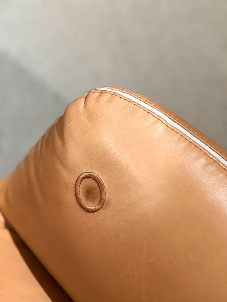 Close-up of tan leather with a circular stitched eyelet and a white seam along the edge.