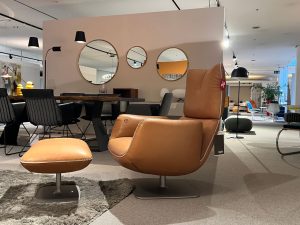 Tan leather swivel lounge chair with matching ottoman in a modern furniture showroom, circular wall mirrors nearby