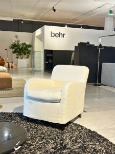 White upholstered armchair on a dark shag rug in a modern showroom with a Behr sign on a white wall in the background.