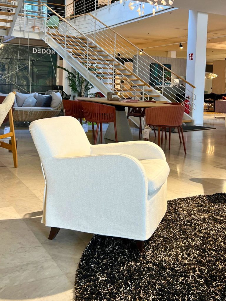 White upholstered armchair on a black shag rug in a modern showroom with a staircase and assorted seating in the background