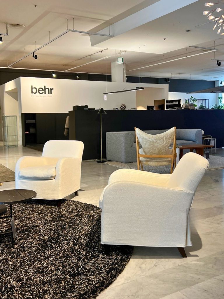 Showroom seating area with white upholstered chairs around a black shag rug; Behr logo on a white wall in the background.