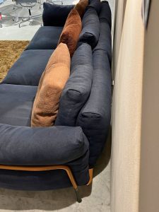 Navy blue sectional sofa with brown and dark blue cushions placed against a wall in a showroom. Visible chairs and a brown rug in the background.
