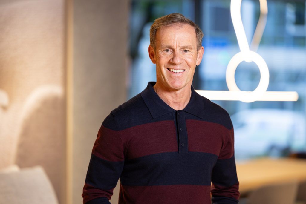 Smiling middle-aged man in a navy and burgundy striped polo posing in a modern office lobby.