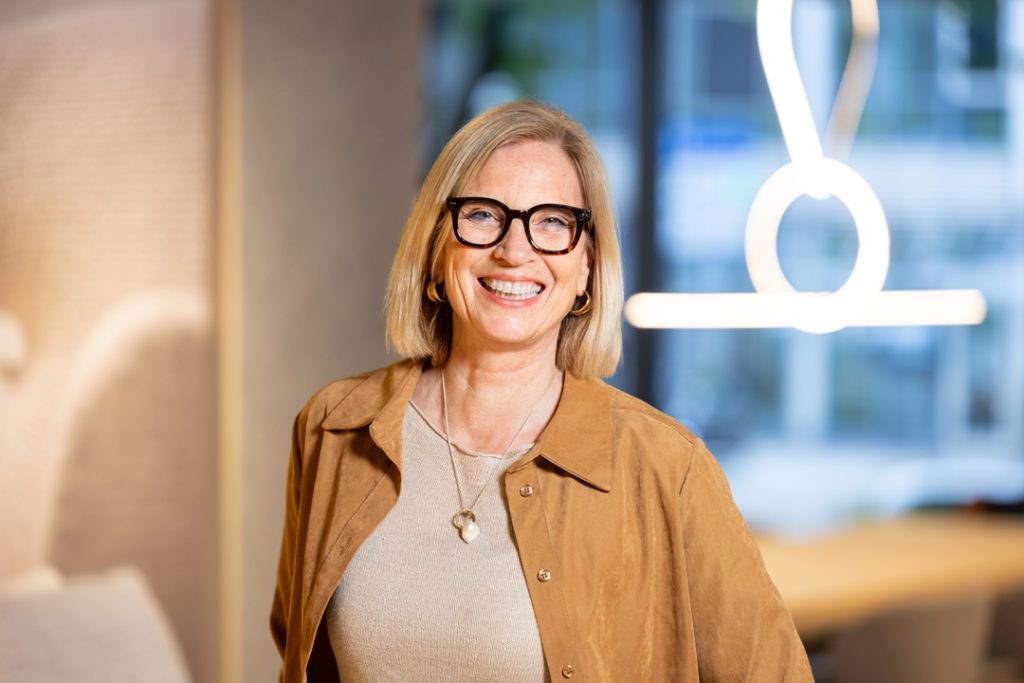 Smiling blonde woman with black glasses in a tan jacket, standing in a modern office setting.
