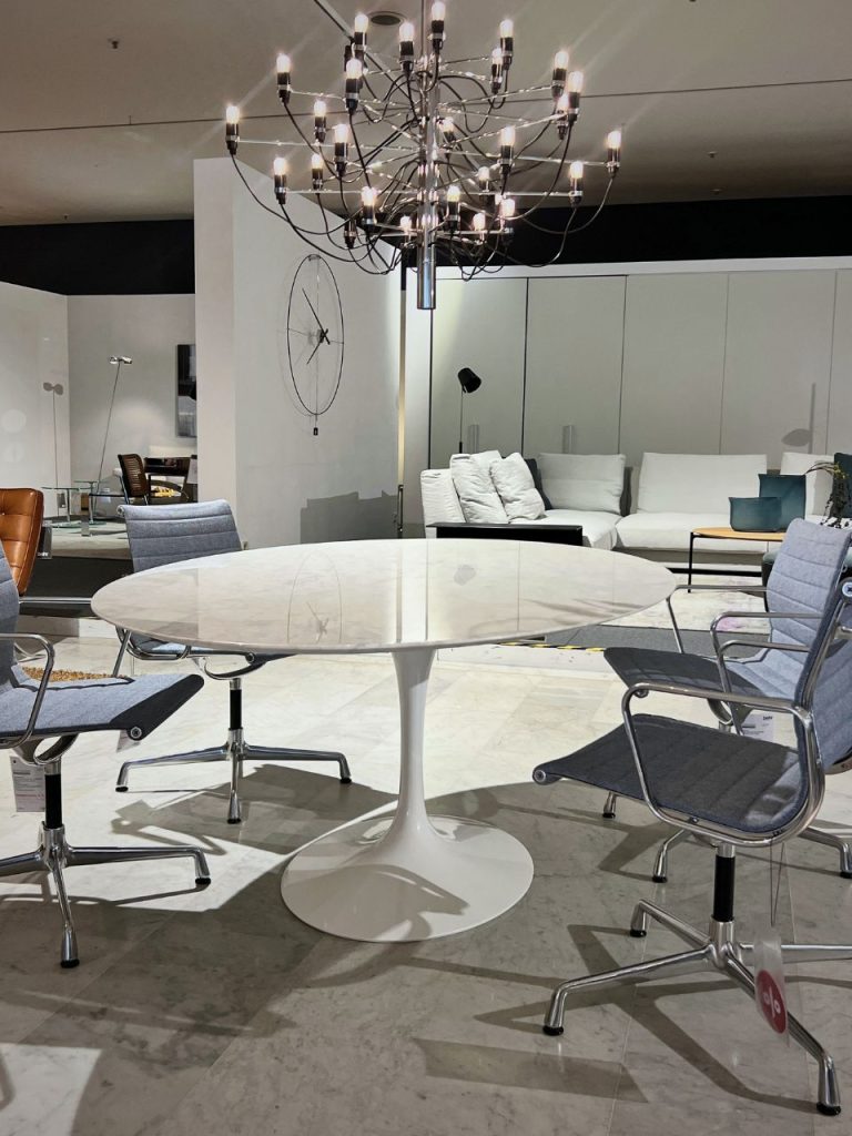 Round white marble pedestal table surrounded by four gray cushioned office chairs in a modern showroom setting.