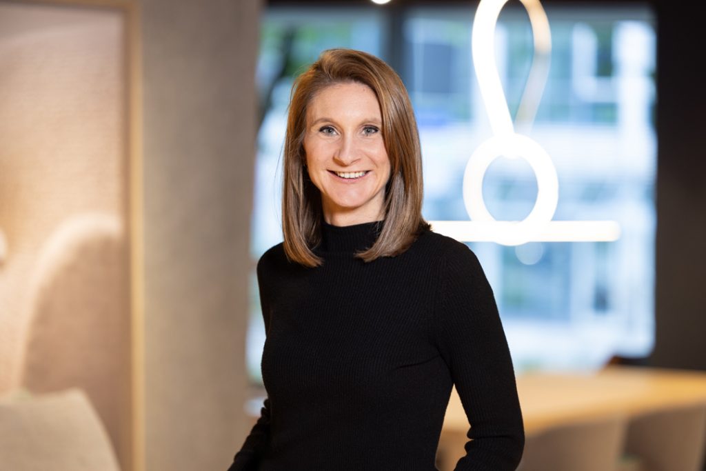 Smiling woman in a black turtleneck stands in a modern office lobby.