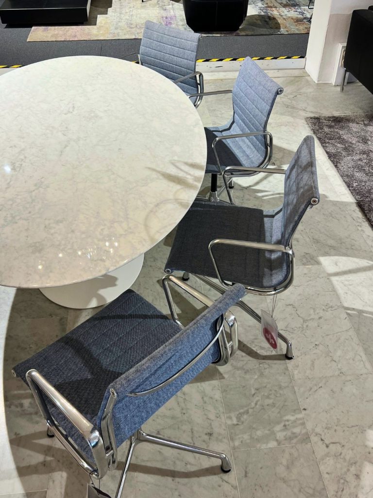 Round white marble table surrounded by four blue upholstered chairs with chrome frames in a modern showroom setting.