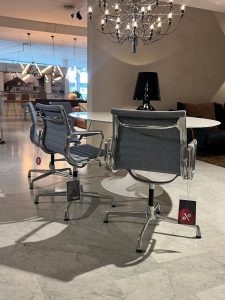 Showroom scene with gray and chrome swivel chairs around a white round table, a black lamp in the center, and a chandelier overhead.