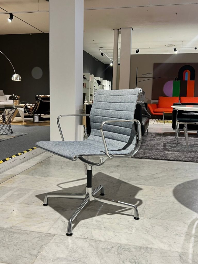 Gray fabric swivel chair with chrome armrests and five-star base on a marble floor in a furniture showroom. In the background are sofas, lamps, and shelves displaying more furniture pieces.