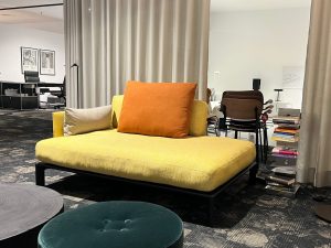 Bright yellow sofa with orange and beige cushions in a modern showroom lounge, with curtains behind and a magazine stack nearby.