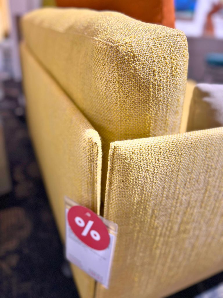 Close-up of yellow textured upholstered furniture (chair arm) with a red circular discount tag showing a percent symbol hanging from it