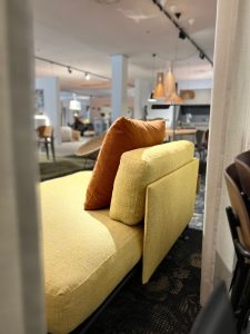 Yellow fabric sofa with an orange cushion in a bright furniture showroom, foreground focus and blurred background signals a showroom setting.