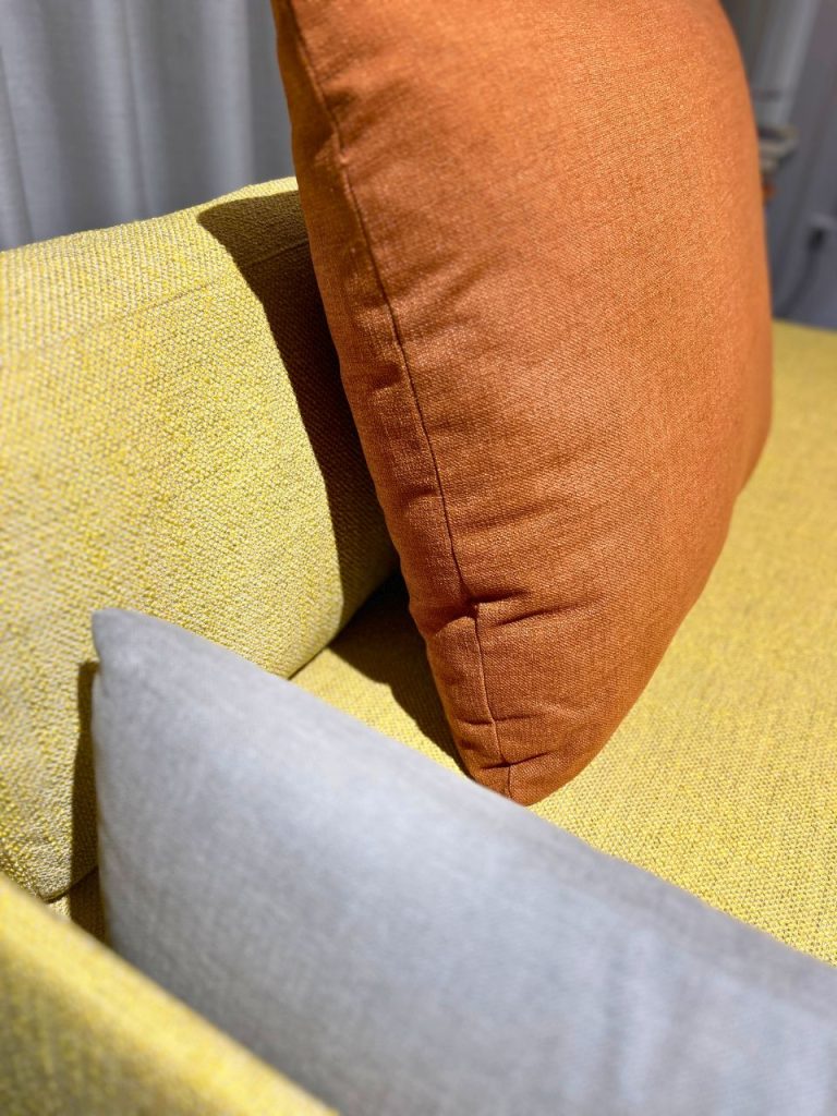 Close-up of a mustard-yellow sofa with a burnt-orange cushion leaning against it and a light gray pillow in the foreground.