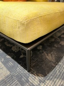 Close-up of a yellow upholstered ottoman with a black metal leg on a dark patterned rug.
