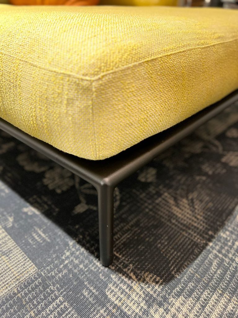 Close-up of a yellow upholstered ottoman with a black metal leg on a dark patterned rug.