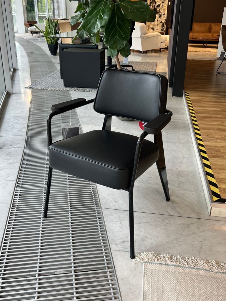 Black office chair with padded seat and backrest in a modern lobby setting near plants and a metal floor grille