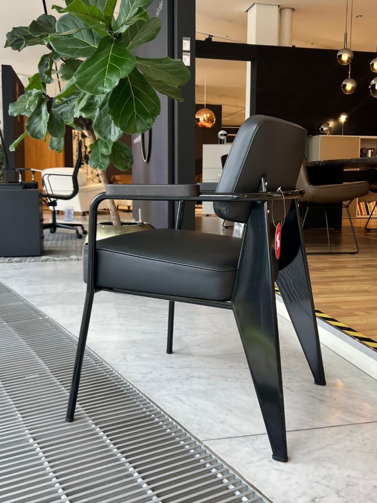 Black contemporary lounge chair with slender metal arms in a furniture showroom; a large potted plant sits nearby and other seating is in the background with warm lighting.