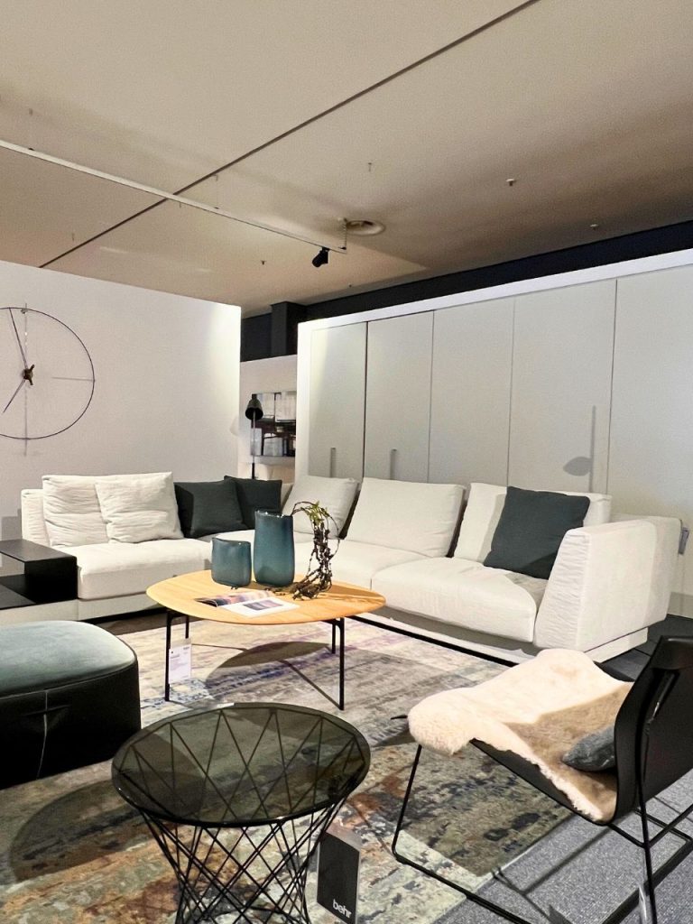 Modern showroom living room with a white sectional sofa, teal cushions, and a round wooden coffee table with blue vases on a patterned rug.