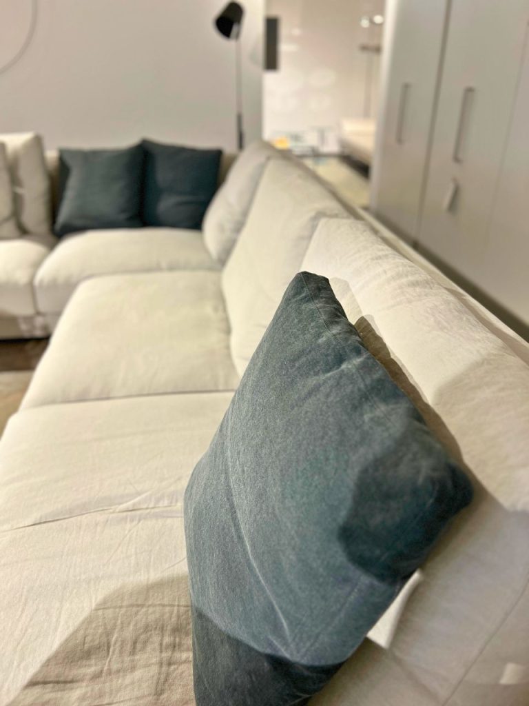 Close-up of a teal throw pillow on a light-colored sectional sofa in a bright, modern living room.
