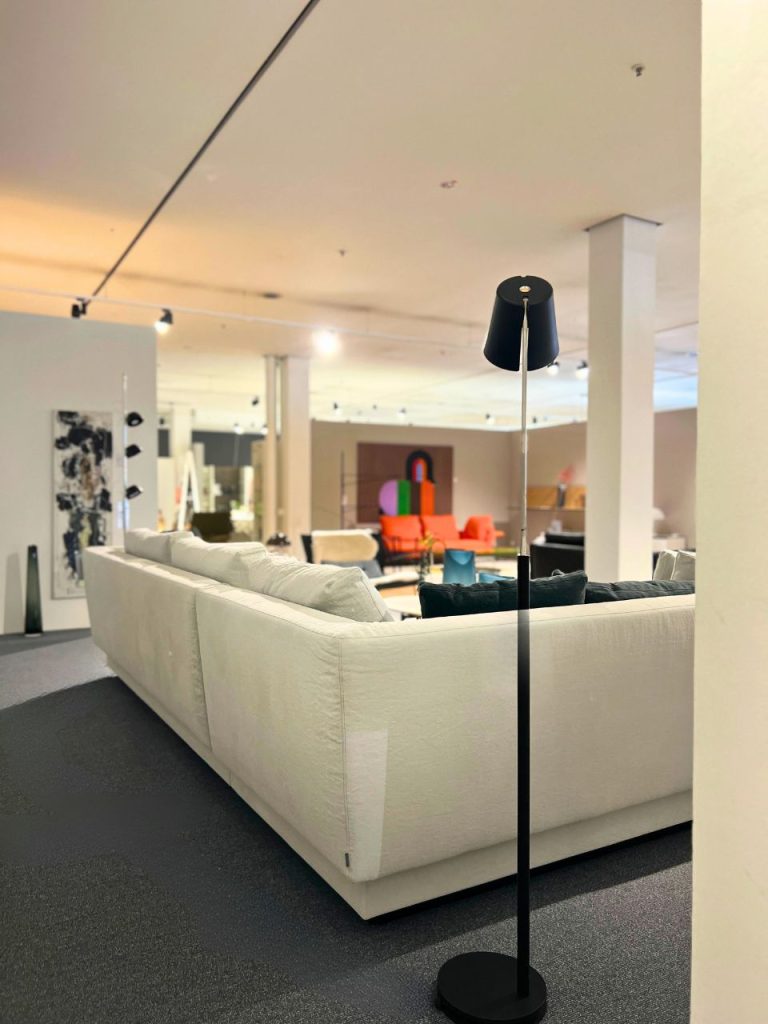 Black floor lamp next to a white sectional sofa in a bright showroom with other colorful furniture in the background.