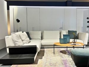White sectional sofa with cream cushions in a modern showroom, a round wooden coffee table with blue glass vases and a magazine in front.