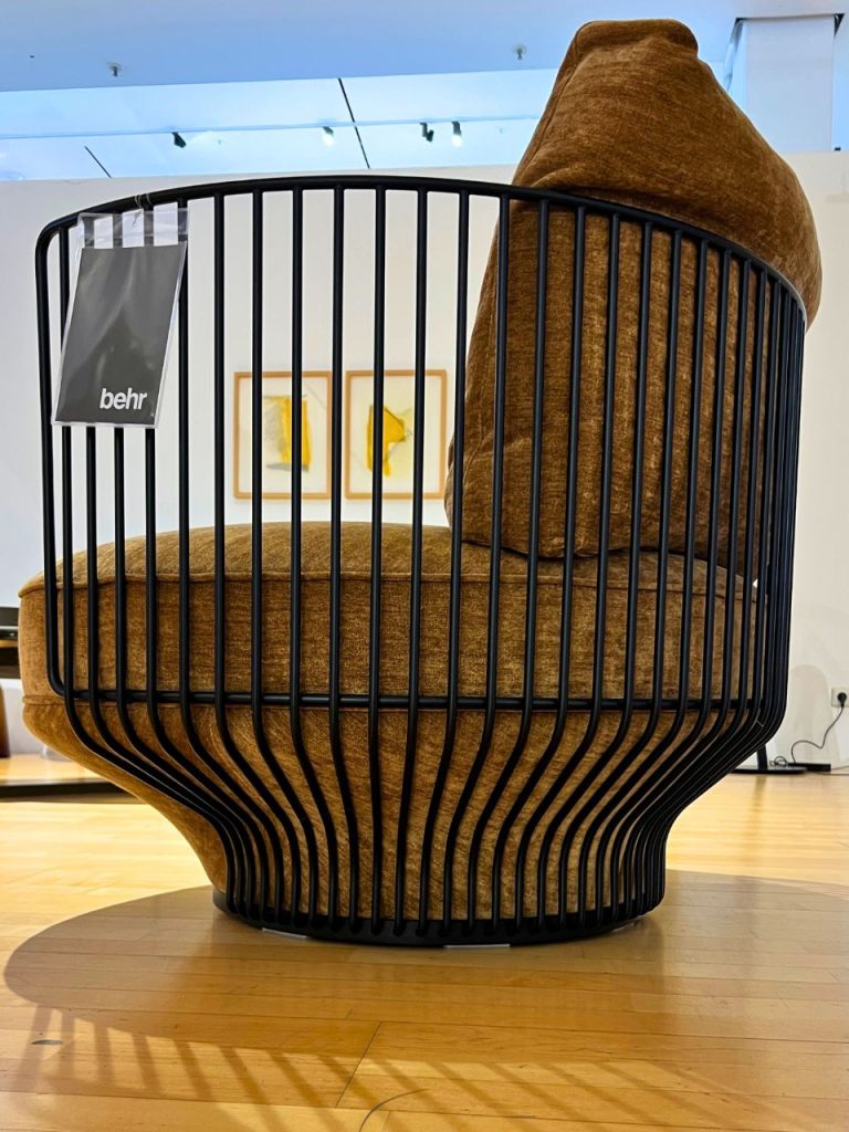 Brown cocoon-style lounge chair with a curved black metal frame, upholstered in brown fabric; a Behr tag hangs from the side, in an art gallery or showroom.