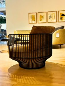 In a furniture showroom, a round black metal-framed lounge chair with beige cushions sits on a wooden floor, with abstract art prints on the wall behind it.
