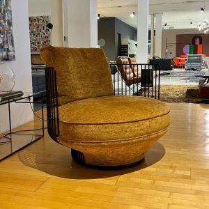 Round mustard-yellow velvet lounge chair with black metal rails, on a wooden showroom floor.