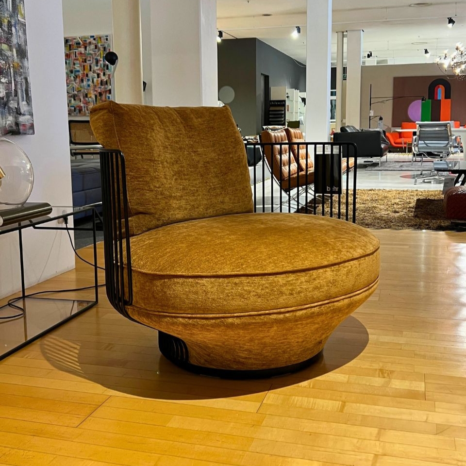 Round mustard-yellow velvet lounge chair with black metal rails, on a wooden showroom floor.
