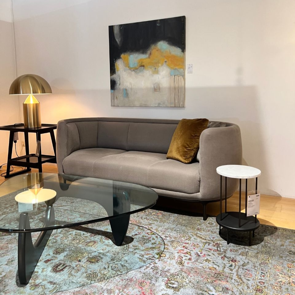 Modern living room setup: curved grey sofa with mustard pillow, glass coffee table, and a brass dome lamp on a side table beside it. Visual rug underfoot and abstract wall art above the sofa.