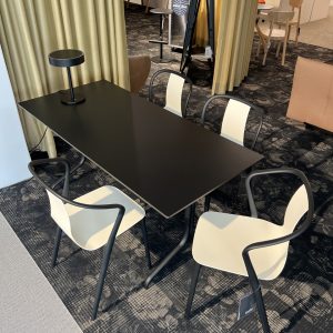 Modern black rectangular table with four white-and-black chairs around, a black lamp on the table, in a showroom setting with patterned carpet and dividers in the background.