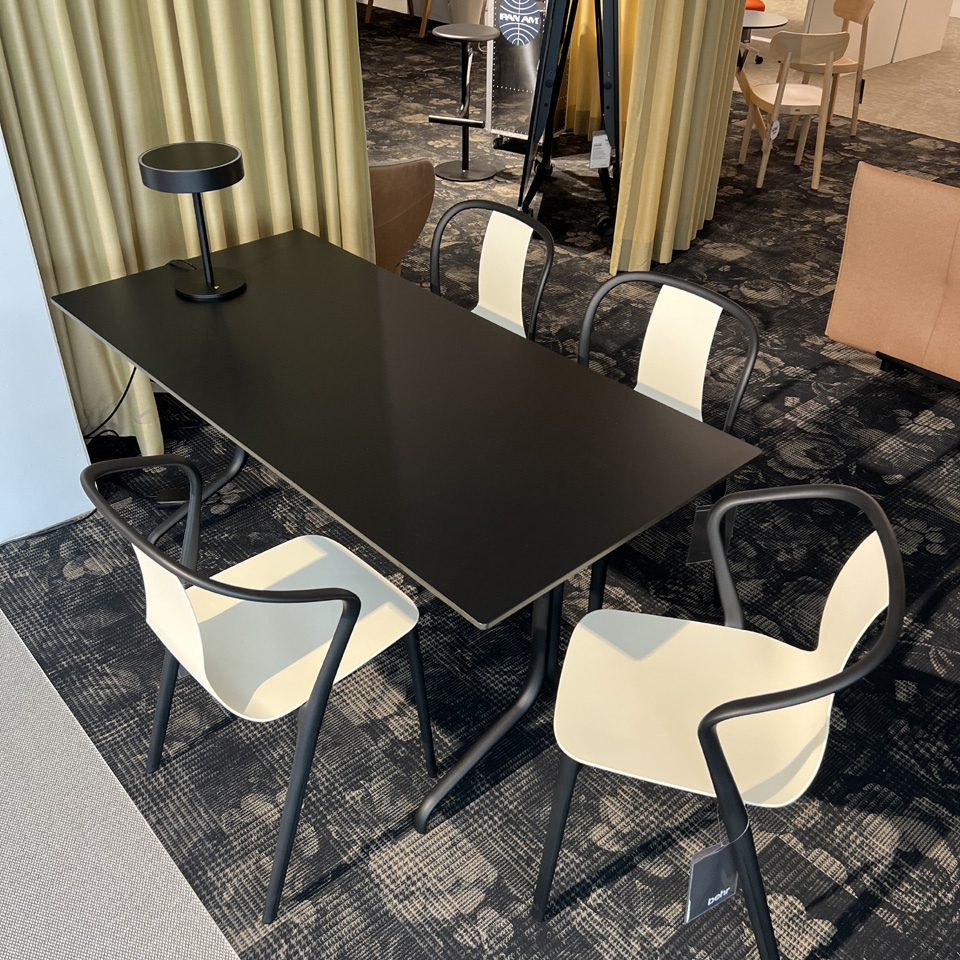 Modern black rectangular table with four white-and-black chairs around, a black lamp on the table, in a showroom setting with patterned carpet and dividers in the background.