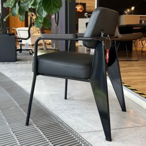 Black metal-framed lounge chair with a padded seat and back, on display in a modern showroom with other chairs and a plant in the background.