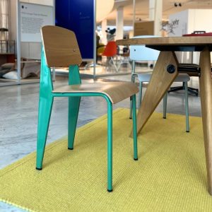 Teal metal-framed chair with a light wood seat and backrest, beside a wooden table on a yellow rug in a showroom.